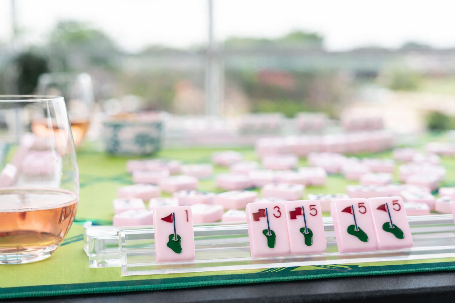 Close-up of a golf-themed mahjong game with pink tiles and a glass of wine on a green surface.