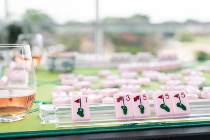 Close-up of a golf-themed mahjong game with pink tiles and a glass of wine on a green surface.
